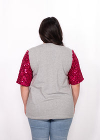 Woman with dark hair standing in a back profile position wearing the Howdy Honey Top against a white background. The focal point of this image is the pink sequin sleeves against grey fabric. The model paired this top with light-wash denim jeans. 