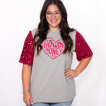 Woman with dark hair standing in a front profile position wearing the Howdy Honey Top against a white background. The focal point of this image is the pink sequin sleeves and pink howdy honey graphic. The model paired this top with light-wash denim jeans. 