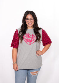 Woman with dark hair standing in a front profile position wearing the Howdy Honey Top against a white background. The focal point of this image is the pink sequin sleeves and pink howdy honey graphic. The model paired this top with light-wash denim jeans. 