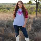 Woman with red hair standing in a front profile position wearing the Howdy Honey Top in a lifestyle scene. The focal point of this image is the Howdy Honey top in a scene with heavy greenery. The model paired this top with dark-wash denim jeans.  