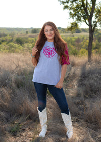 Woman with red hair standing in a front profile position wearing the Howdy Honey Top in a lifestyle scene. The focal point of this image is the Howdy Honey top in a scene with heavy greenery. The model paired this top with dark-wash denim jeans.  