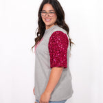 Woman with dark hair standing in a side profile position wearing the Howdy Honey Top against a white background. The focal point of this image is the pink sequin sleeve. The model paired this top with light-wash denim jeans. 
