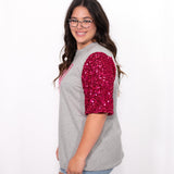 Woman with dark hair standing in a side profile position wearing the Howdy Honey Top against a white background. The focal point of this image is the pink sequin sleeve. The model paired this top with light-wash denim jeans. 