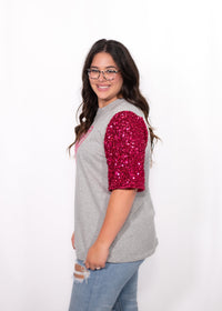 Woman with dark hair standing in a side profile position wearing the Howdy Honey Top against a white background. The focal point of this image is the pink sequin sleeve. The model paired this top with light-wash denim jeans. 