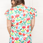 Woman with dark hair standing in a back profile position wearing the Iris Top against a white background. The focal point of this image is the colorful floral graphics on back of top. The model paired this top with dark-wash denim shorts. 