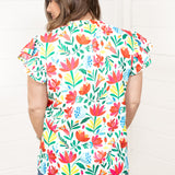 Woman with dark hair standing in a back profile position wearing the Iris Top against a white background. The focal point of this image is the colorful floral graphics on back of top. The model paired this top with dark-wash denim shorts. 
