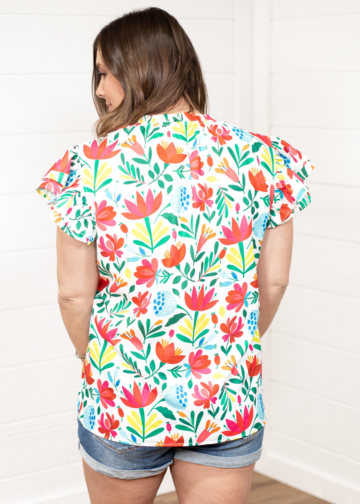 Woman with dark hair standing in a back profile position wearing the Iris Top against a white background. The focal point of this image is the colorful floral graphics on back of top. The model paired this top with dark-wash denim shorts. 