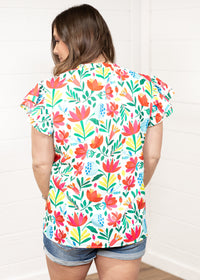 Woman with dark hair standing in a back profile position wearing the Iris Top against a white background. The focal point of this image is the colorful floral graphics on back of top. The model paired this top with dark-wash denim shorts. 