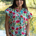 Woman with dark hair standing in a front profile position wearing the Iris Top in a lifestyle scene. The focal point of this image is front floral graphics, v-neckline, and ruffle sleeves. The model paired this top with light-wash denim jeans. 