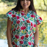 Woman with dark hair standing in a front profile position wearing the Iris Top in a lifestyle scene. The focal point of this image is front floral graphics, v-neckline, and ruffle sleeves. The model paired this top with light-wash denim jeans. 