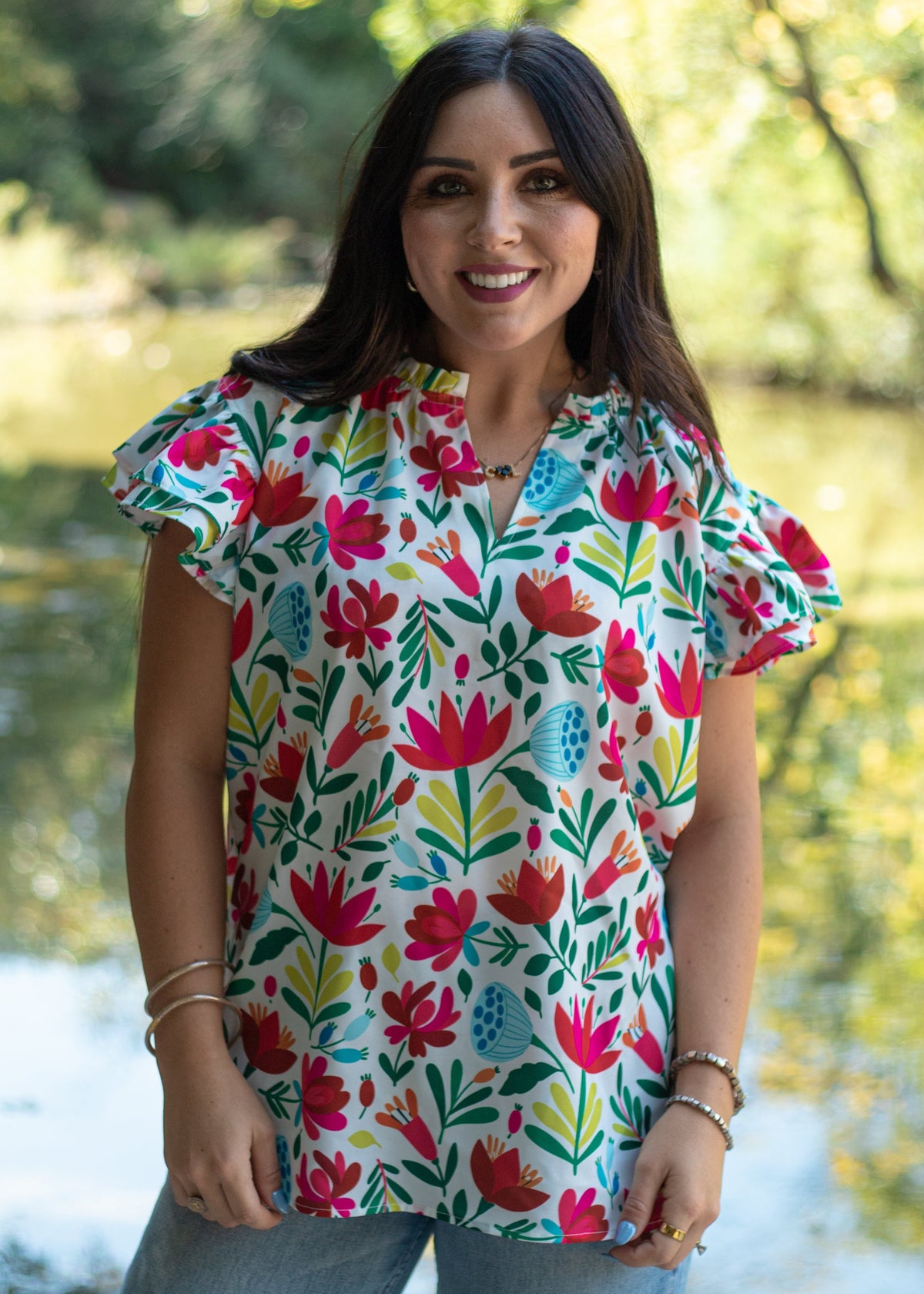 Woman with dark hair standing in a front profile position wearing the Iris Top in a lifestyle scene. The focal point of this image is front floral graphics, v-neckline, and ruffle sleeves. The model paired this top with light-wash denim jeans. 