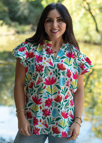 Woman with dark hair standing in a front profile position wearing the Iris Top in a lifestyle scene. The focal point of this image is front floral graphics, v-neckline, and ruffle sleeves. The model paired this top with light-wash denim jeans. 