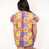 Woman with dark hair standing in a back profile position wearing the Iris Top against a white background. The focal point of the image is the graphics on the back of the top with an emphasis on the ruffle sleeves. The model paired this top with dark-wash denim shorts.