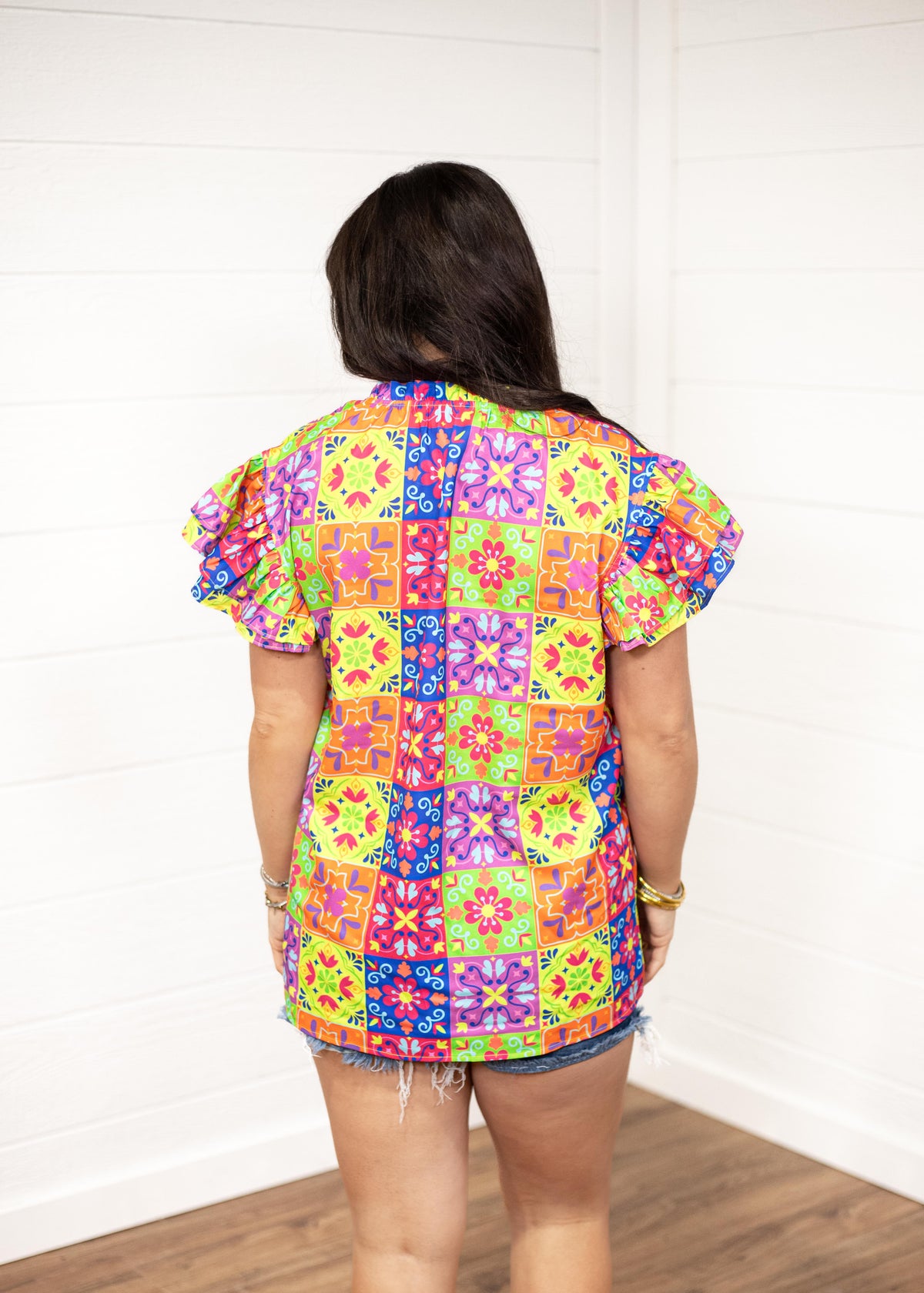 Woman with dark hair standing in a back profile position wearing the Iris Top against a white background. The focal point of the image is the graphics on the back of the top with an emphasis on the ruffle sleeves. The model paired this top with dark-wash denim shorts.