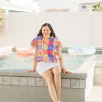 Woman with dark hair sitting in front profile position wearing the Iris Top against a "real time" background with a pool. The focal point of this image is the top in it's entirety. The model paired this top with a white skirt and silver sneakers. 