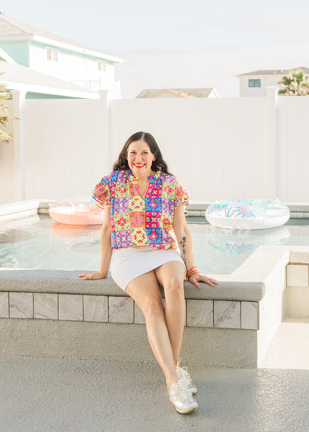 Woman with dark hair sitting in front profile position wearing the Iris Top against a "real time" background with a pool. The focal point of this image is the top in it's entirety. The model paired this top with a white skirt and silver sneakers. 