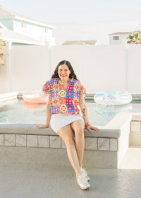 Woman with dark hair sitting in front profile position wearing the Iris Top against a "real time" background with a pool. The focal point of this image is the top in it's entirety. The model paired this top with a white skirt and silver sneakers. 