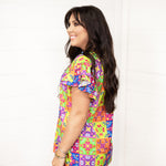 Woman with dark hair standing in a side profile position wearing the Iris Top against a white background. The focal point of this image is the ruffle sleeves and graphics on the side of the top. The model paired this top with dark-wash denim shorts. 