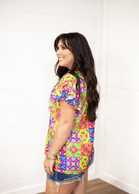 Woman with dark hair standing in a side profile position wearing the Iris Top against a white background. The focal point of this image is the ruffle sleeves and graphics on the side of the top. The model paired this top with dark-wash denim shorts. 