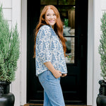 Woman with red hair wearing a quarter-sleeve top with blue Santa toile graphics and blue embroidery on shoulders and chest. Model paired this top with denim jeans.