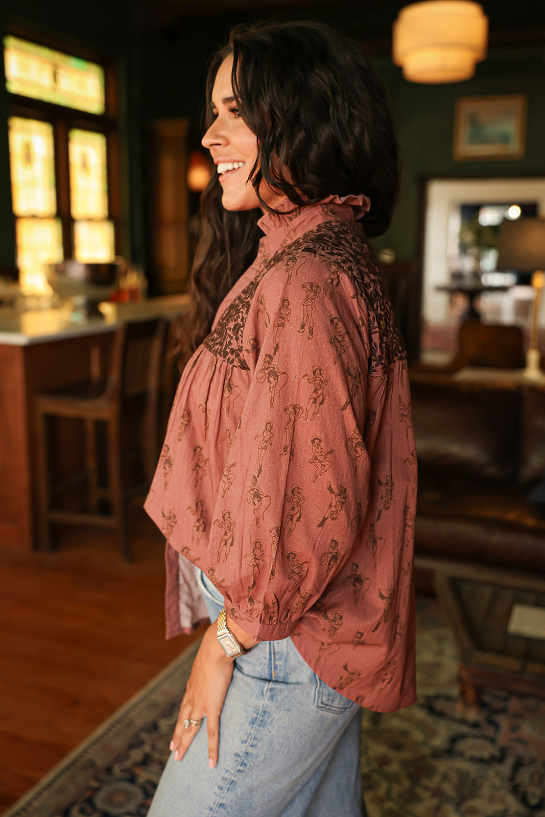 Woman with dark hair standing in a side profile position wearing the Jane Top in a lifestyle scene. The focal point of this image is the cowgirl graphics as well as embroidery along shoulders. The model paired this top with light-wash denim jeans. 