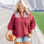 Woman with blonde hair standing in a front profile position wearing the Jane Top at a football field. The focal point of this image is the embroidery on the shoulders, buttons down the front of the top, and the v-neckline. The model paired this top with light-wash denim shorts.