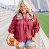 Woman with blonde hair standing in a front profile position wearing the Jane Top at a football field. The focal point of this image is the embroidery on the shoulders, buttons down the front of the top, and the v-neckline. The model paired this top with light-wash denim shorts.