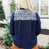 Woman with blonde hair wearing blue quarter sleeve blouse with white embroidery on front and back. and a burnt orange skirt.