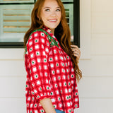 Woman with red hair wearing a quarter-sleeve top with red plaid graphics and green embroidery. Model paired this top with light-wash denim jeans. 