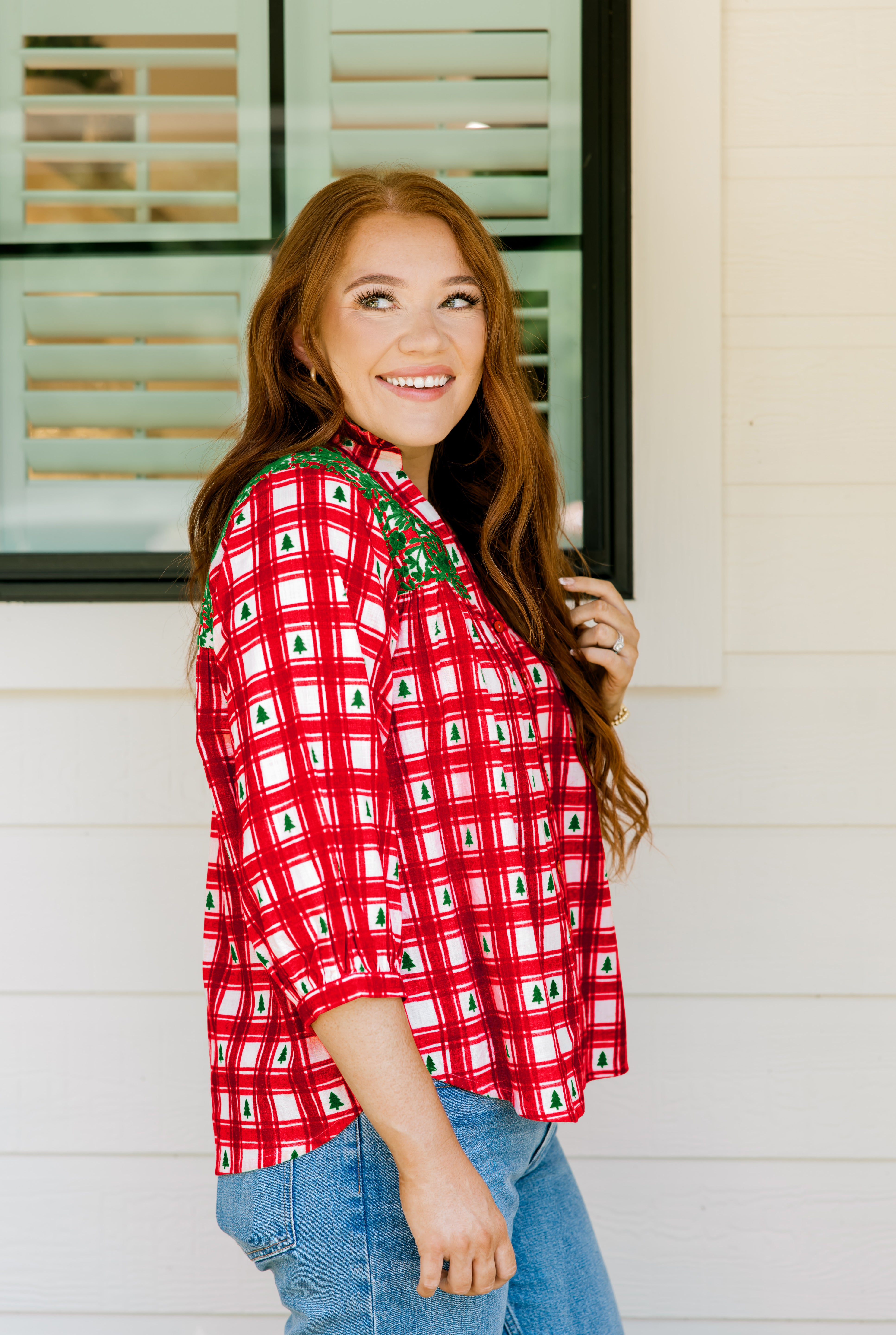Woman with red hair wearing a quarter-sleeve top with red plaid graphics and green embroidery. Model paired this top with light-wash denim jeans. 