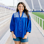 Woman with dark hair standing in a front profile position wearing the Jane Top at a football field. The focal point of this image is the embroidery on both shoulders, buttons down the front of top, high-collar and v-neckline. The model paired this top with dark-wash denim shorts. 