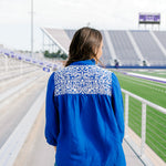 Woman with dark hair is standing in a back profile position wearing the Jane Top at a football field. The focal point of this image is the white embroidery on the shoulders and pleated back. The model paired dark-wash denim shorts with this top. 