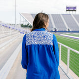 Woman with dark hair is standing in a back profile position wearing the Jane Top at a football field. The focal point of this image is the white embroidery on the shoulders and pleated back. The model paired dark-wash denim shorts with this top. 