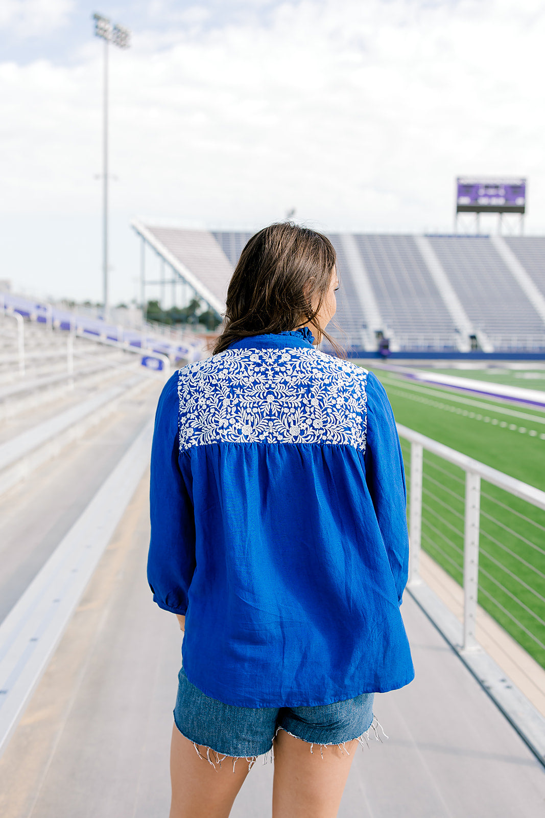 Woman with dark hair is standing in a back profile position wearing the Jane Top at a football field. The focal point of this image is the white embroidery on the shoulders and pleated back. The model paired dark-wash denim shorts with this top. 