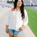 Woman with dark hair standing in a front profile position wearing the Jane Top at a football field. The focal point of this image is the buttons down the front of the top, the burnt orange embroidery as well as the v-neckline. The model paired this top with light-wash denim shorts.