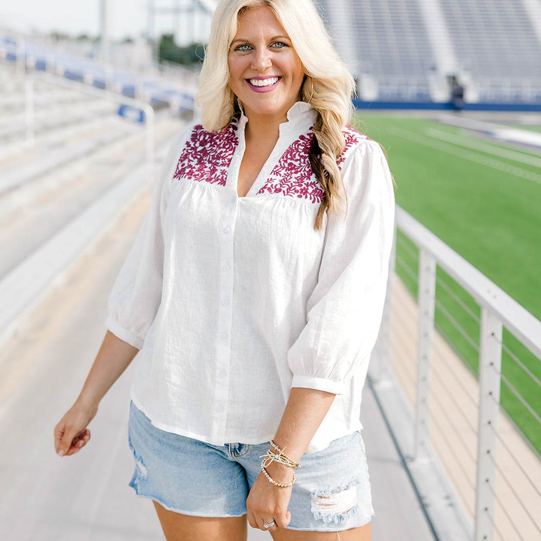 Woman with blonde hair standing in a front profile position wearing the Jane Top at a football field. The focal point of this image is the embroidery across front of top, buttons down the front of shirt and the v-neckline with a high collar. The model paired this top with light-wash denim shorts.