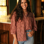 Woman with dark hair standing in a front profile position wearing the Jane Top in a lifestyle scene. The focal point of this image is the cowgirl graphics, buttons down front of top and embroidery on shoulders. The model paired this top with light-wash denim jeans. 