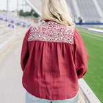 Woman with blonde hair standing in a back profile position wearing the Jane Top at a football field. The focal point of this image is the embroidery across the shoulders and pleating beneath. The model paired this top with light-wash denim shorts.