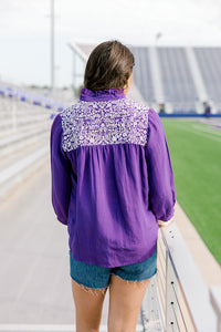 Woman with dark hair standing back profile wearing the Jane Top at a football field. The focal point of this image is the embroidery along the shoulders, pleated back, and high collar. The model paired this top with dark-wash denim shorts.