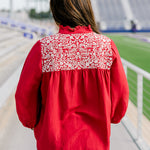 Woman with dark hair standing in a back profile position wearing the Jane Top at a football field. The focal point of this image is the white embroidery across the shoulders, pleating on back as well as the high collar with ruffle. The model paired this top with light-wash denim shorts.