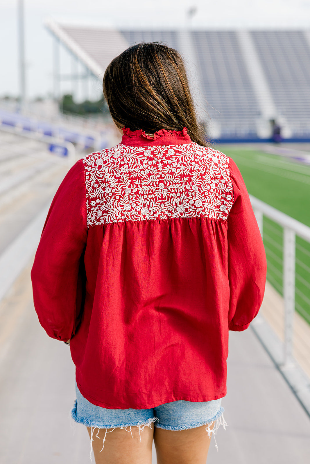 Woman with dark hair standing in a back profile position wearing the Jane Top at a football field. The focal point of this image is the white embroidery across the shoulders, pleating on back as well as the high collar with ruffle. The model paired this top with light-wash denim shorts.