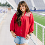 Woman with dark hair standing in a front profile position wearing the Jane Top at a football field. The focal point of this image is the buttons down the front of top, pleating beneath embroidery on shoulders, and quarter sleeves with pleating towards cuff. The model paired this top with light-wash denim shorts.