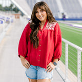 Woman with dark hair standing in a front profile position wearing the Jane Top at a football field. The focal point of this image is the buttons down the front of top, pleating beneath embroidery on shoulders, and quarter sleeves with pleating towards cuff. The model paired this top with light-wash denim shorts.