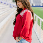 Woman with dark hair standing in a side profile position wearing the Jane Top at a football field. The focal point of this image is the quarter sleeve with pleating to the cuff as well as embroidery over the shoulders. The model paired this top with light-wash denim shorts. 
