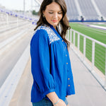 Woman with dark hair standing in the side profile position wearing the Jane Top at a football field. The focal point of this image is the quarter sleeve that is slightly pleated at the cuff, embroidery on shoulder and sneak peek of the button down in the front. The model paired this top with dark-wash shorts.