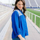 Woman with dark hair standing in the side profile position wearing the Jane Top at a football field. The focal point of this image is the quarter sleeve that is slightly pleated at the cuff, embroidery on shoulder and sneak peek of the button down in the front. The model paired this top with dark-wash shorts.
