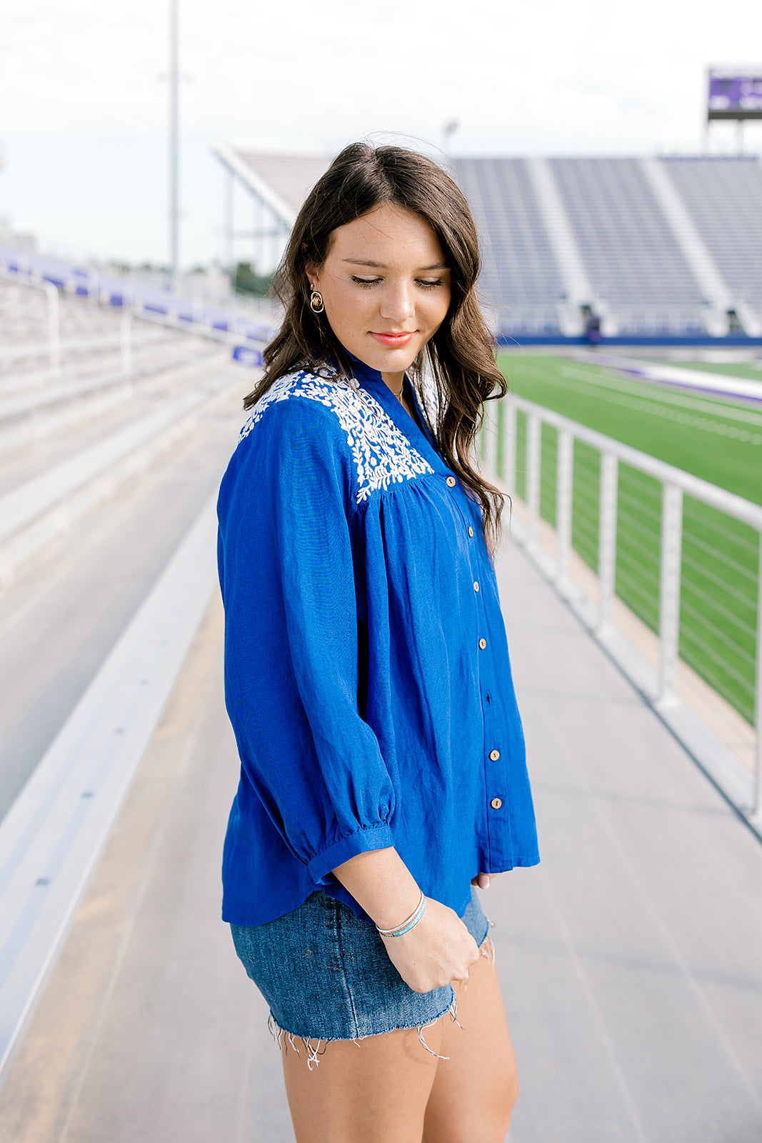Woman with dark hair standing in the side profile position wearing the Jane Top at a football field. The focal point of this image is the quarter sleeve that is slightly pleated at the cuff, embroidery on shoulder and sneak peek of the button down in the front. The model paired this top with dark-wash shorts.
