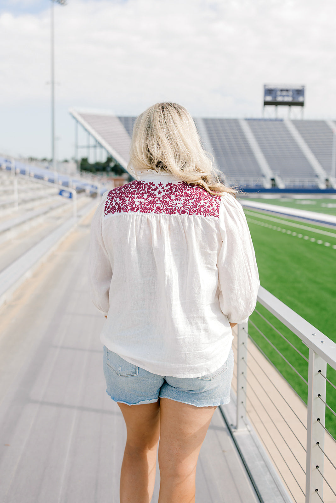 Woman with blonde hair standing in a back profile position wearing the Jane Top at a football field. The focal point of this image is the embroidery across the shoulders, pleating beneath, as well as emphasis on the ruffled collar. The model paired this top with light-wash denim shorts.