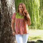 Woman with red hair standing front profile in lifestyle scene that has heavy greenery in Jeanie Top. She is wearing the Jeanie Top paired with white pants. 
