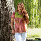 Woman with red hair standing front profile in lifestyle scene that has heavy greenery in Jeanie Top. She is wearing the Jeanie Top paired with white pants. 